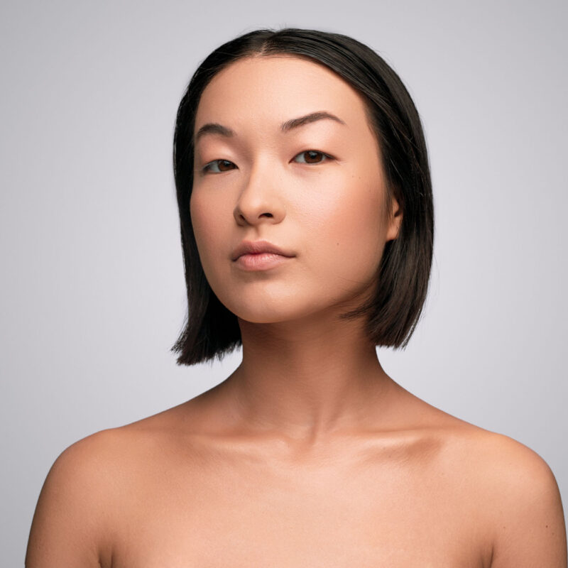 Attractive ethnic female with clean skin looking at camera during spa session against gray background